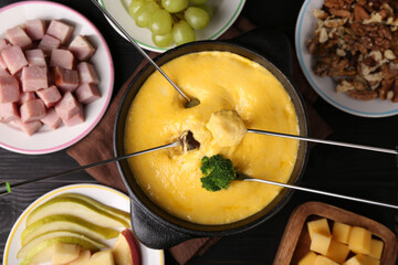 Dipping different products into fondue pot with melted cheese on black wooden table, flat lay