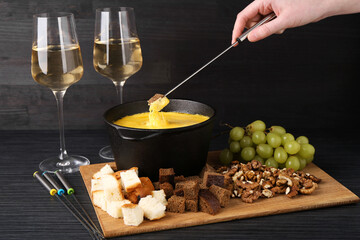 Woman dipping piece of bread into fondue pot with melted cheese at black wooden table, closeup