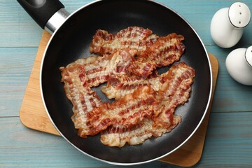 Delicious bacon slices in frying pan on blue wooden table, flat lay