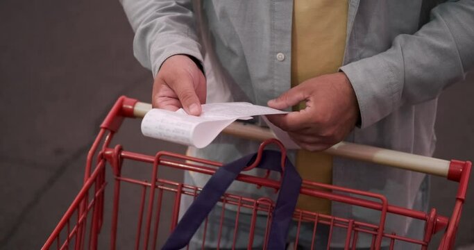 View of the hands of a man standing next to a grocery cart and holding a receipt with a list of products and expenses. Counting money spent at the grocery store. Saving money.