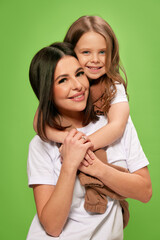 Happy, beautiful, smiling young woman, mother hugging with cute, tender little girl, daughter against green studio background. Concept of happiness, Mother's day, childhood, fashion and lifestyle