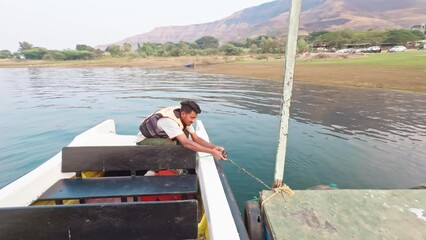 smiling young Man in a life vest sitting on a yacht moor the boat ashore by a sea or river with mountains in the background