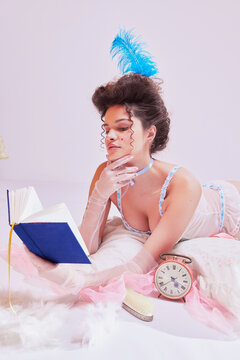 Attractive young woman in tender lingerie with feathered headpiece lying on bed with book. Vintage boudoir aesthetics. Historical romance novels. Concept of beauty and fashion, vintage, boudoir style