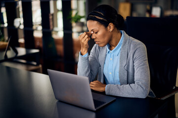 A tired African woman has a headache while working online, at the office.
