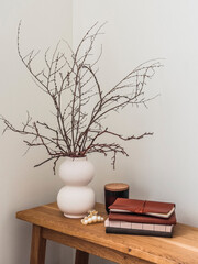 Ceramic vase with dry branches, books, scented candle on a wooden table in the living room