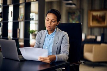 A focused woman doing paperwork, at the office while using a laptop.