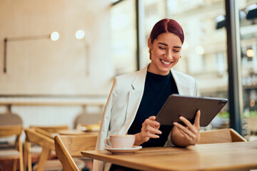 A smiling red-haired beautiful woman, using a tablet while sitting at the cafe and drinking coffee.