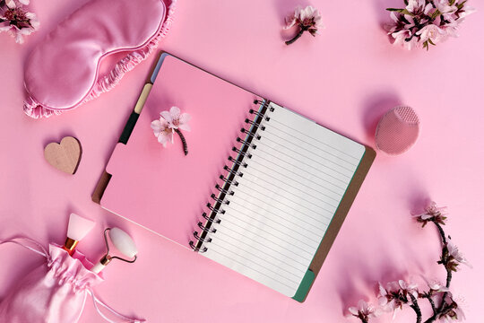 Square pink overhead background, quality of sleep, and self care routine. Sleeping mask, blank notebook with copy-space, massage stone roller, cream and wooden heart on pink paper.