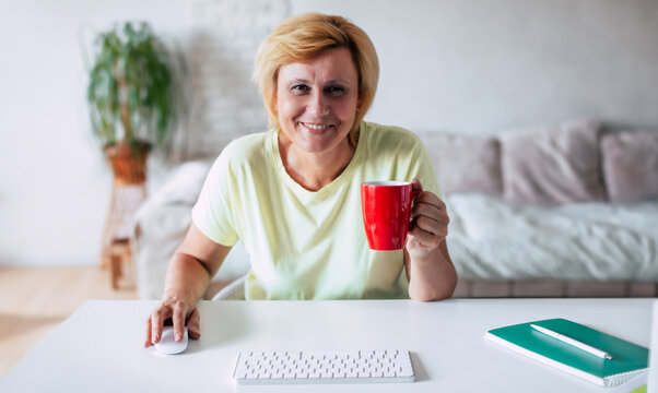 Close up shot of middle-aged beautiful smiling woman in casual clothes using a computer with keypad and mouse at home. Mature woman is drinking coffee or tea