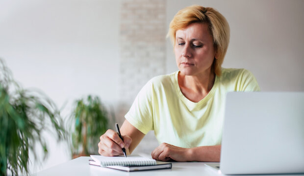 Close up image of concentrated mature woman while she is writing with pen on notepad at home