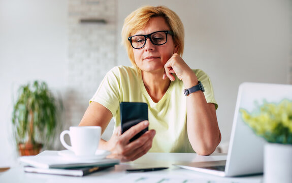 Close up portrait of happy mature woman using mobile phone while working at home with laptop. Smiling female wearing eyeglasses messaging or chatting in phone. Lady browsing site on cellphone