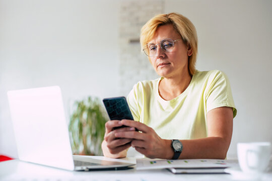  Side view image of middle aged woman with smart phone in hands during online shopping. Surfing in internet. Female wearing eyeglasses chatting in phone during work at home office