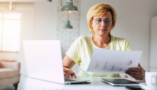 Photo of concentrated mature woman in glasses working with modern laptop and reading report from work documents in her apartment