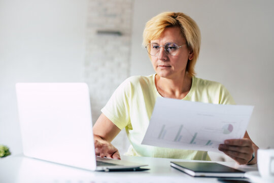 Middle aged woman sit with laptop and paper document, pensive  mature lady reading paper bill pay online at home managing bank finances calculating taxes planning loan debt pension payment