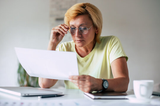 Close up portrait of clever middle-aged woman in goggles with papers while sitting by workplace in front of laptop at home office