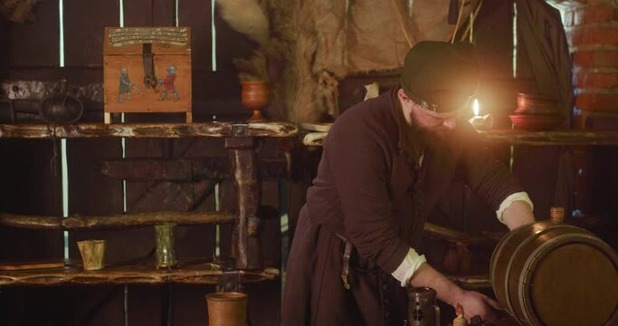 Barkeeper pours ale from barrel and turns to counter at inn. Plump bartender in medieval suit looks with servile smile at guest in tavern. Fantasy world