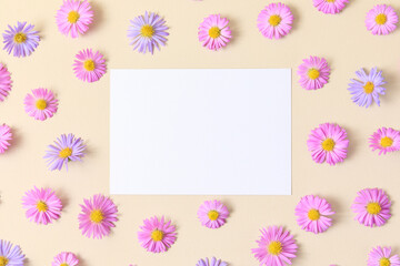 Greeting card template. Pink and purple flower aster on beige table background with blank paper sheet. Flat lay, top view, mockup, copy space for text. Aesthetic stylish floral pattern.