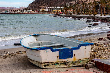 Old rowboat on sand on coastal beach with sea, boardwalk and marina in background, waves crashing on rocks, cloudy day in La Paz, Baja California Sur Mexico