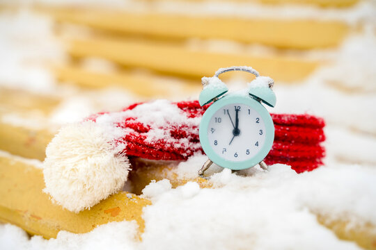 A small alarm clock on a yellow snow-covered bench in a winter snow park. Concept of winter, Christmas, new year, warm, cozy, loving, protecting
