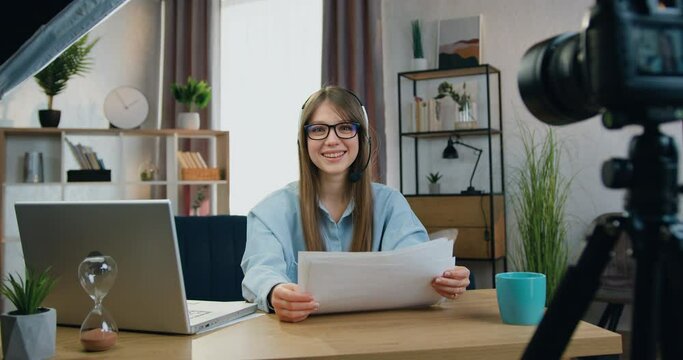 Smiling young woman in headphones sitting in front of camera before the beginning of online stream with prepared documents about her work