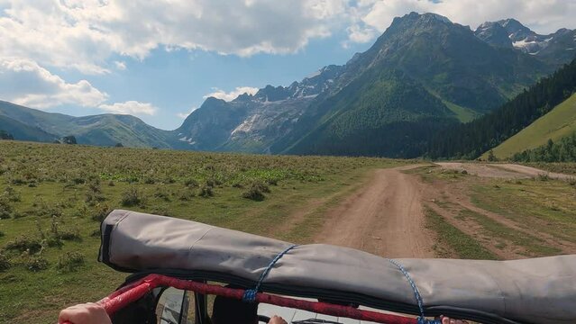 SUV driving on a dirt road in the mountains. First person view of a man standing in the car with the roof open. High mountains with glaciers ahead. Summer off-road driving. Slow motion
