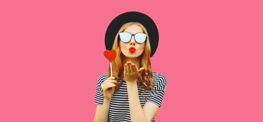 Portrait of beautiful young woman blowing her lips sends kiss with sweet red heart shaped lollipop