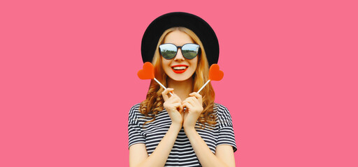 Portrait of happy cheerful young woman with sweet red heart shaped lollipop on stick on pink