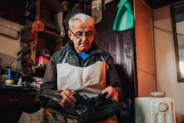 A senior craftsman is sitting in his shop and repairing boots.