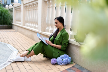 Stylish adult woman wearing green costume, sitting on city street near architectural columns with hydrangea flowers bouquet, reading magazine - Powered by Adobe