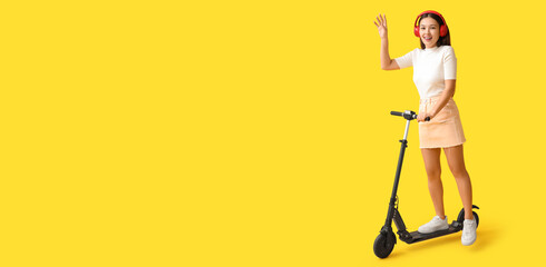 Beautiful young woman in headphones riding modern electric kick scooter on yellow background with space for text