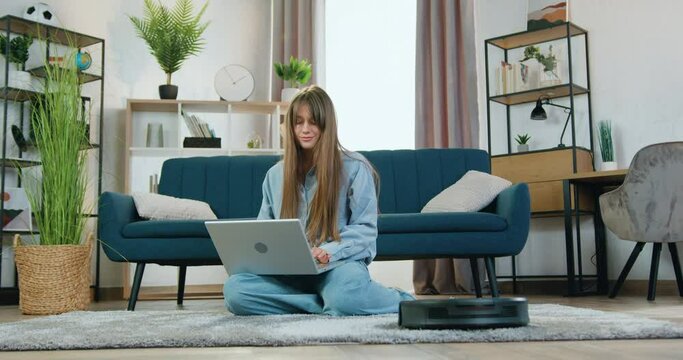 Portrait of woman which resting on carpet at home and listening music on laptop while automatic vacuum cleaner cleaning carpet