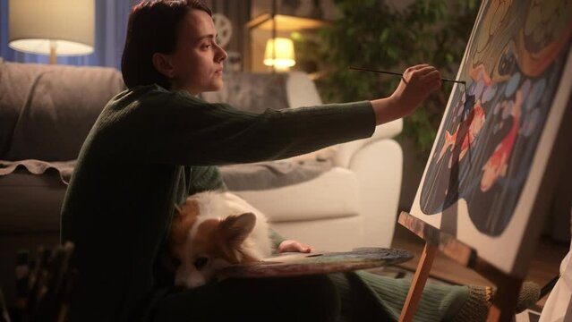 A woman is petting her fluffy corgi dog while creating her work of art. Inspired girl illustrator working at home. A beautiful young female artist is painting a picture in her cozy studio
