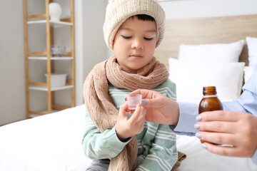 Ill little Asian boy taking cough syrup from his mother in bedroom