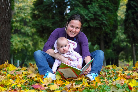 Portrait of happy young mother and little baby relaxing in park. Mom reading a fairytale to her daughter. Horizontal image.