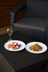 there is food on the rattan table. Pilaf and Greek vegetable salad and a glass of white wine on the table. Eat dinner outside. Garden furniture for home. rattan furniture on the terrace