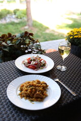 there is food on the rattan table. Pilaf and Greek vegetable salad and a glass of white wine on the table. Eat dinner outside. Garden furniture for home. rattan furniture on the terrace
