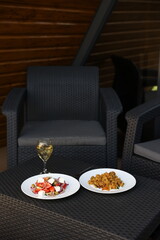 there is food on the rattan table. Pilaf and Greek vegetable salad and a glass of white wine on the table. Eat dinner outside. Garden furniture for home. rattan furniture on the terrace