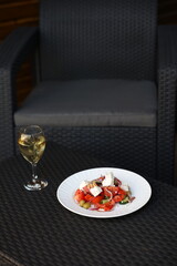 there is food on the rattan table. Greek vegetable salad and a glass of white wine on the table. Eat dinner outside. Garden furniture for home. rattan furniture on the terrace