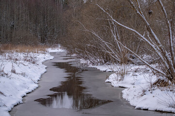 winter landscape with small forest river. Water partially covered with ice. Calm stream in quiet evening 