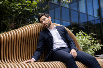 Thoughtful businessman in suit sitting on outdoor bench by modern office