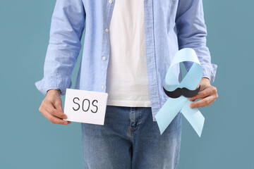 Young man holding paper with word SOS and blue ribbon on color background, closeup. Prostate cancer awareness concept