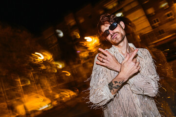 An edgy young man in sequined attire strikes a pose under the night lights, his tattoos adding to his cool, carefree nightlife vibe.