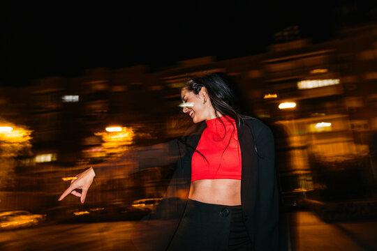 Laughing young woman with dark hair enjoys a vibrant night scene with dynamic light painting effects, pointing playfully.