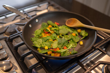 Frying pan sauteeing fresh vegetables with a wooden spoon on a gas range.