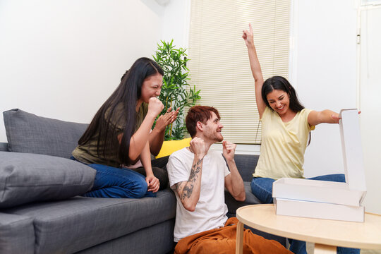 Three friends express triumph and joy in their apartment, celebrating with pizza. A man and two women cheer by the couch.