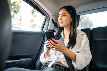 Fototapeta na wymiar Asian Woman Traveler sitting in car back seats using smartphone call taxi service in application. Online app with cell phone and put on safety belt. Female passenger in car traveling to destination.