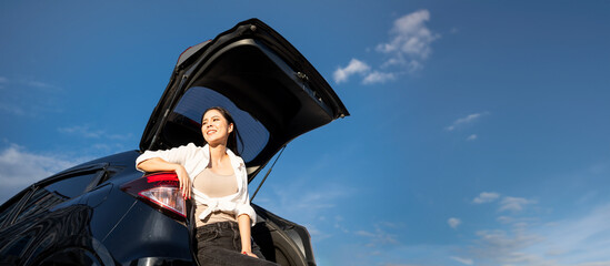 Young beautiful asian women buying new car. she was sitting at trunk of car. Beautiful moment blue sky Smiling female driving travel by vehicle on the road on a bright day weekend with sun light.