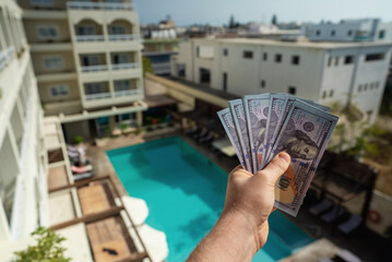 Man holds hundred dollar bills in front of a hotel with a swimming pool.