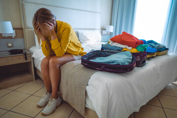 Flight cancellation. Upset woman sitting on bed with suitcases with clothes. - obrazy, fototapety, plakaty