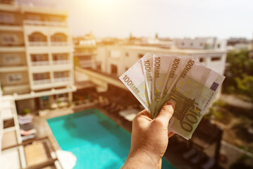 Man holds hundred-euro bills in front of a hotel with a swimming pool.